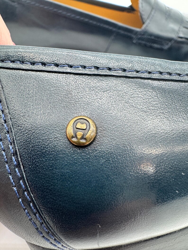May include: Close-up of a navy blue leather loafer. The shoe features a gold-colored emblem and blue stitching details. The leather has a textured appearance, and the stitching is visible along the seams.