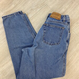 May include: Light blue denim jeans with a classic five-pocket design. The jeans feature a straight leg cut and a leather patch with the brand name "Gitano" on the back. The jeans are folded on a light wood surface.