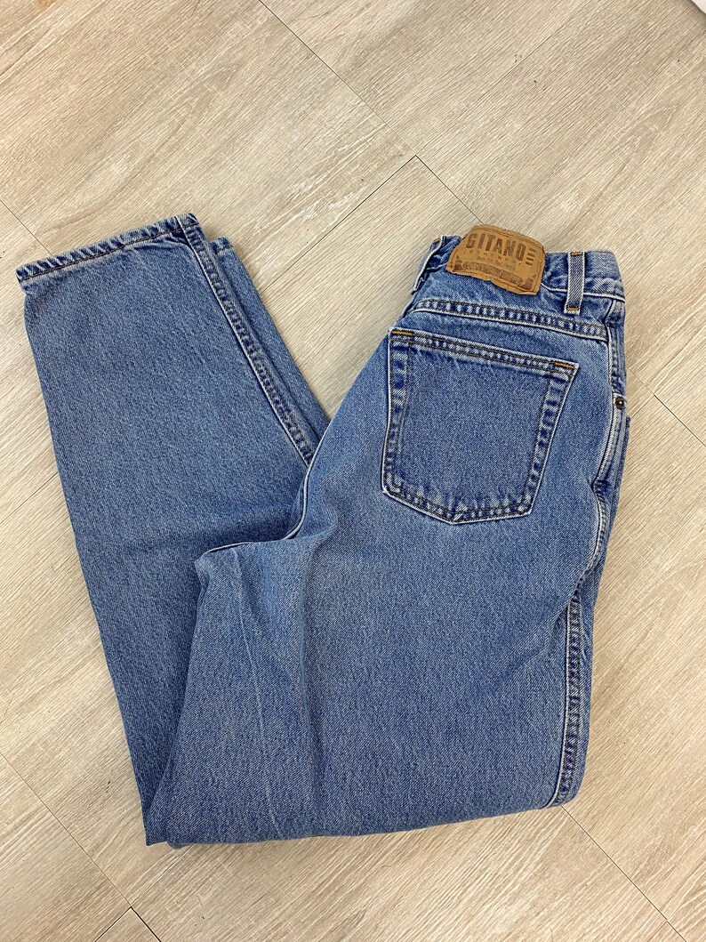 May include: Light blue denim jeans with a classic five-pocket design. The jeans feature a straight leg cut and a leather patch with the brand name "Gitano" on the back. The jeans are folded on a light wood surface.