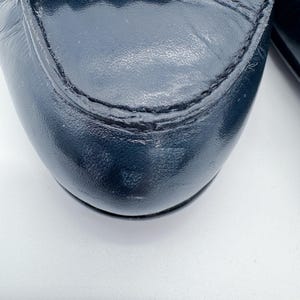 May include: Close-up of a navy blue leather loafer. The shoe features a rounded toe, decorative fringe, and visible stitching. The leather has a slight sheen, and the shoe appears to be well-worn.
