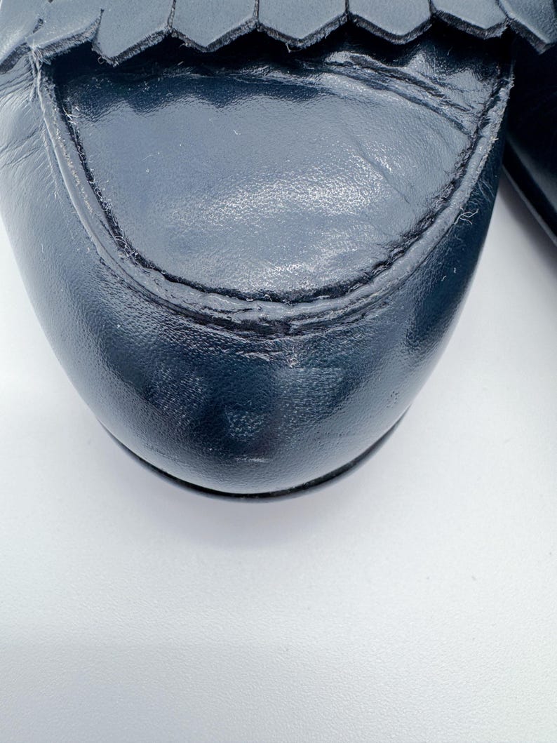 May include: Close-up of a navy blue leather loafer. The shoe features a rounded toe, decorative fringe, and visible stitching. The leather has a slight sheen, and the shoe appears to be well-worn.