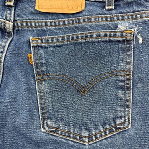 May include: Close-up of a pair of blue denim jeans. The back pocket is visible, with a classic curved stitch design and a small orange tag. The jeans have a worn look with some fraying at the pocket edge.