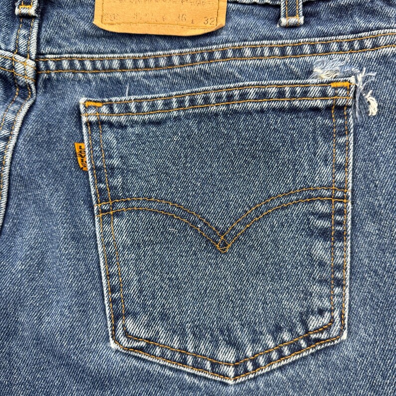 May include: Close-up of a pair of blue denim jeans. The back pocket is visible, with a classic curved stitch design and a small orange tag. The jeans have a worn look with some fraying at the pocket edge.