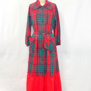May include: A long-sleeved, floor-length dress featuring a red and green plaid pattern. The garment includes a pointed collar, a front button closure, two front patch pockets, a matching fabric waist tie, and a solid red ruffled hemline.