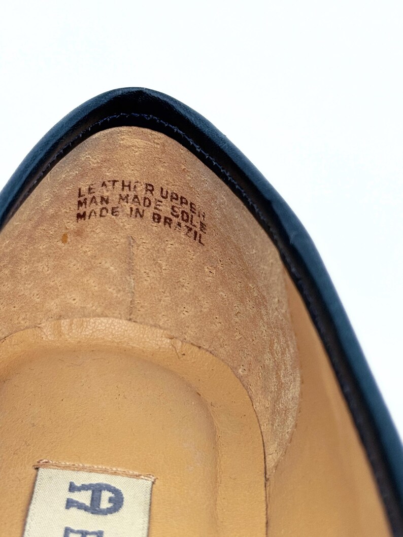 May include: Close-up of the interior of a navy blue shoe. The shoe's insole is tan, with the text "LEATHER UPPER MAN MADE SOLE MADE IN BRAZIL" printed on it. The shoe has a black trim.