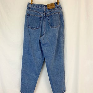 May include: Light blue denim jeans with a high waist, two back pockets, and a leather patch with the brand name "GITANO". The jeans are hanging from a clothesline with wooden clothespins.