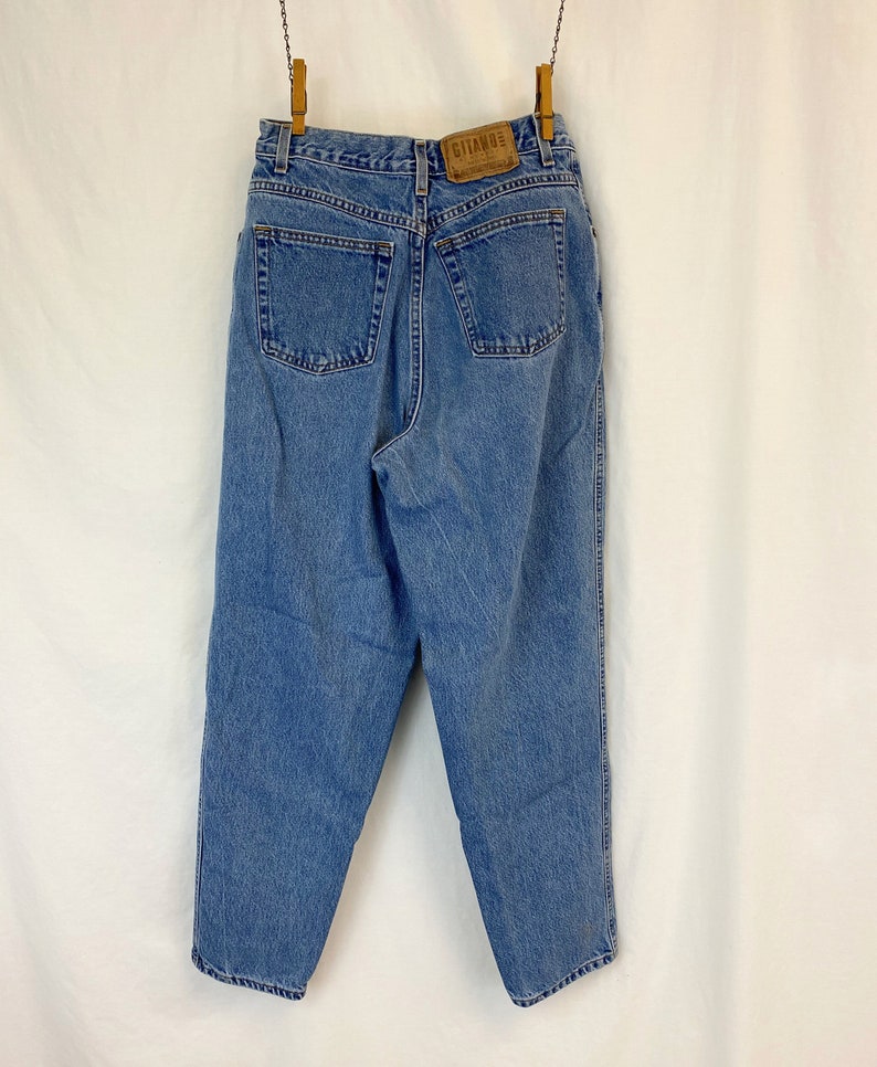 May include: Light blue denim jeans with a high waist, two back pockets, and a leather patch with the brand name "GITANO". The jeans are hanging from a clothesline with wooden clothespins.