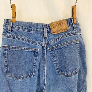 May include: Light blue denim jeans with a classic five-pocket design, hanging from wooden clothespins. The jeans feature a leather patch with the brand name "Gitano". The back pockets and stitching are visible.
