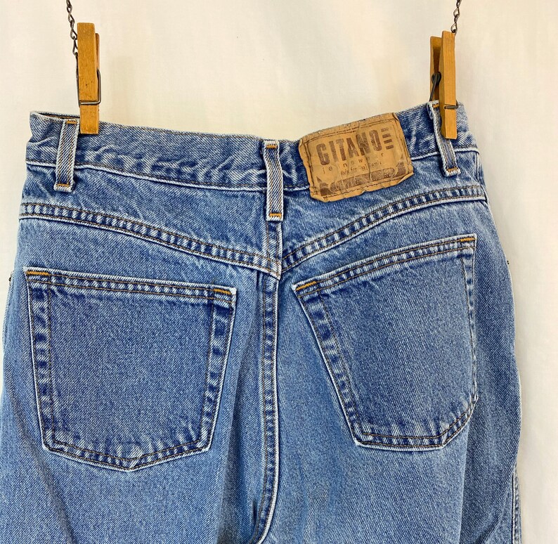 May include: Light blue denim jeans with a classic five-pocket design, hanging from wooden clothespins. The jeans feature a leather patch with the brand name "Gitano". The back pockets and stitching are visible.