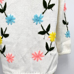 K&ouml;nnte beinhalten: Wei&szlig;er Strickpullover mit gestickten Blumen in den Farben Rosa, Gelb, Blau und Gr&uuml;n.