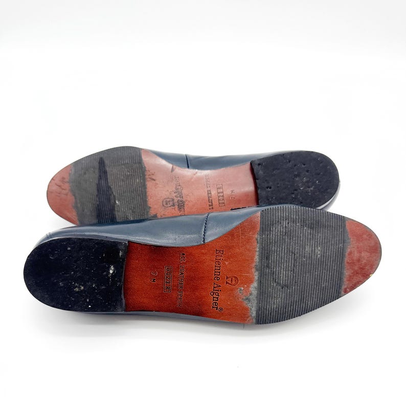 May include: A pair of navy blue leather flats with worn red soles. The soles are marked with the brand name "Etienne Aigner" and size information. The heels are black and show wear.
