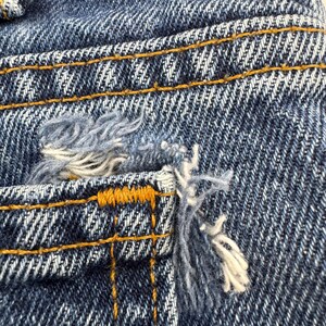 May include: Close-up of distressed denim fabric. The blue jeans show wear and tear with frayed edges and a ripped pocket. The stitching is visible in a contrasting yellow-orange thread.
