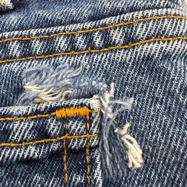 May include: Close-up of distressed denim fabric. The blue jeans show wear and tear with frayed edges and a ripped pocket. The stitching is visible in a contrasting yellow-orange thread.