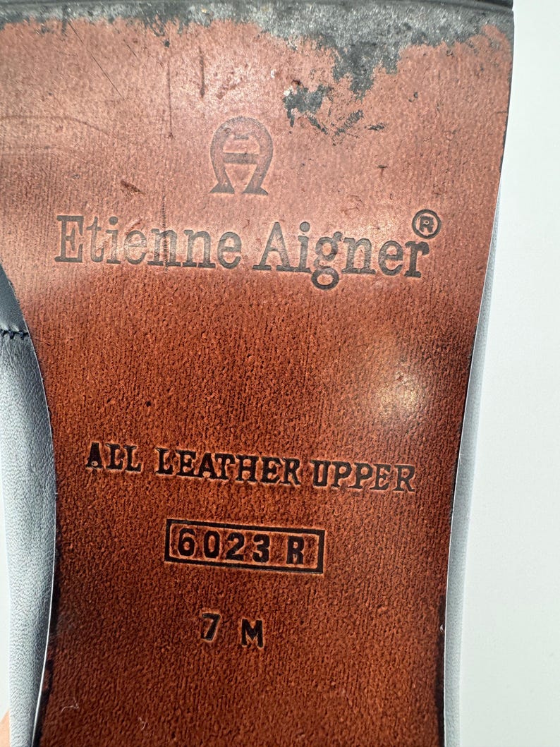 May include: Close-up of the sole of a shoe with the brand name "Etienne Aigner" embossed in the leather. The text "ALL LEATHER UPPER" is visible, along with the size "7 M" and style number "6023 R". The sole is brown.