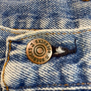 Pu&ograve; includere: Primo piano di una tasca di jeans blu con un bottone color rame che recita "Lee" e "ROBUCKS" in un design circolare.