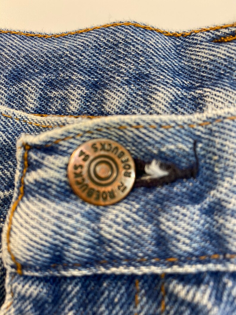 Pu&ograve; includere: Primo piano di una tasca di jeans blu con un bottone color rame che recita "Lee" e "ROBUCKS" in un design circolare.