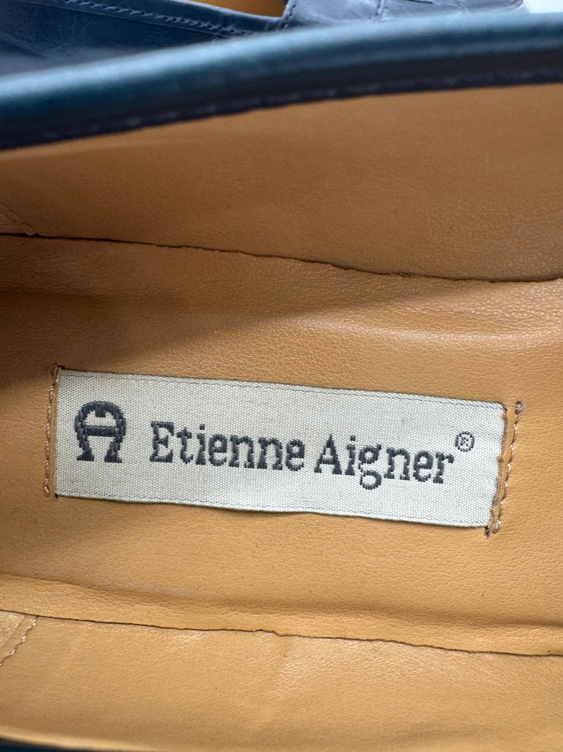 May include: Close-up of the interior of a shoe, featuring a tan leather insole. A rectangular label with the text "Etienne Aigner" is visible. The label has the brand's logo, a stylized "A" with a horseshoe shape.