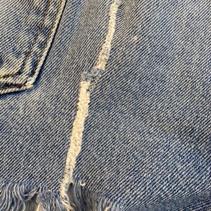 Pu&ograve; includere: Primo piano di un tessuto denim blu chiaro con un bordo sfrangiato e una cucitura bianca che corre al centro.