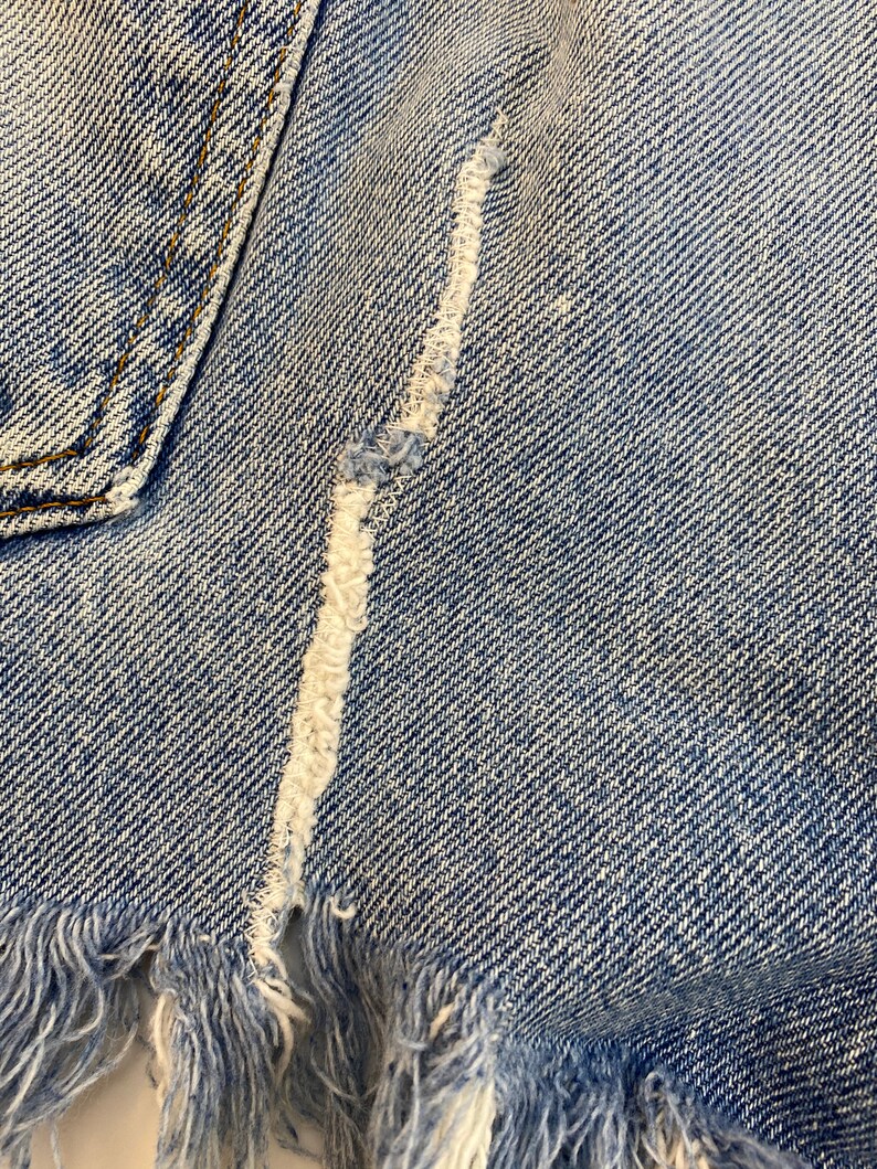 Pu&ograve; includere: Primo piano di un tessuto denim blu chiaro con un bordo sfrangiato e una cucitura bianca che corre al centro.