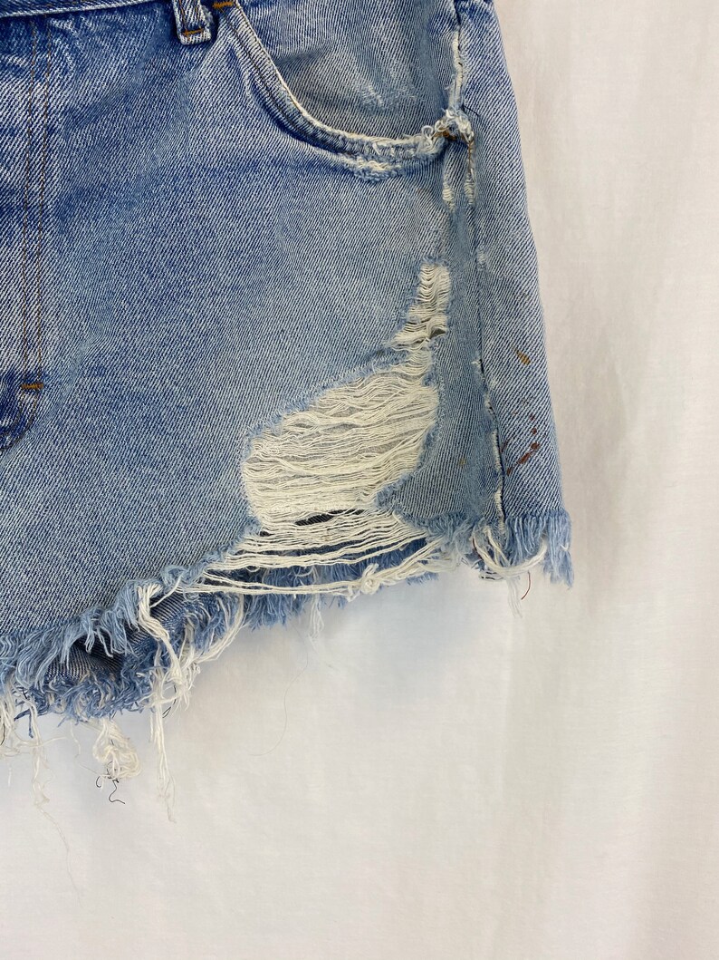 Pu&ograve; includere: Shorts di jeans blu chiaro con bordi sfrangiati e un aspetto consumato. I pantaloncini hanno una tasca anteriore e alcuni piccoli buchi.