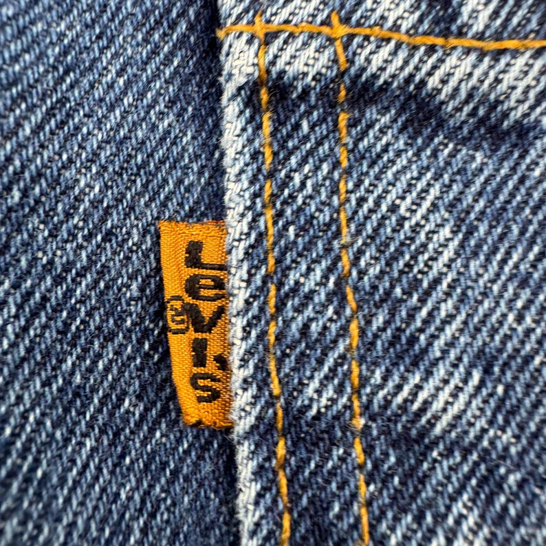 May include: Close-up of a section of blue denim jeans. An orange tag with the word "Levi's" in black lettering is sewn into the jeans. The stitching is in a contrasting yellow-orange color.