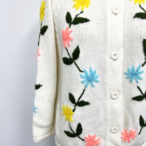 K&ouml;nnte beinhalten: Ein wei&szlig;er Strickcardigan mit gestickten Blumen in Rosa-, Blau- und Gelbt&ouml;nen. Der Cardigan hat einen Knopfschlie&szlig;e.