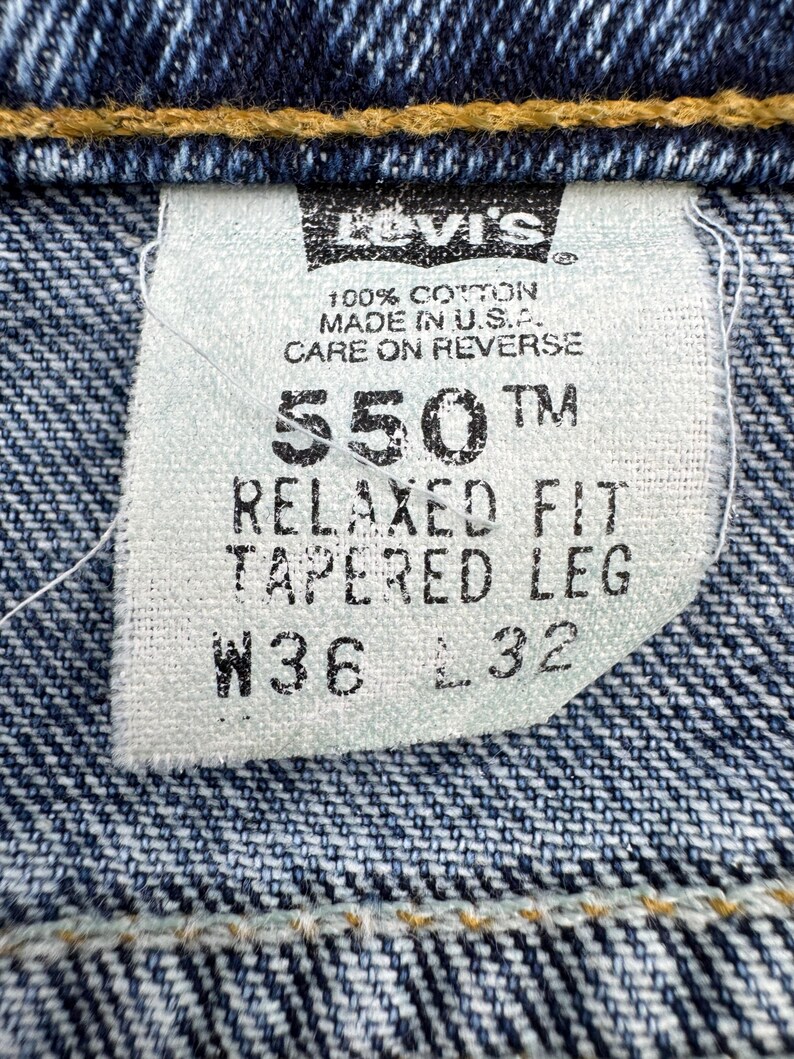 May include: Close-up of a Levi's jeans tag. The tag reads "550 TM RELAXED FIT TAPERED LEG W36 L32" and "100% Cotton Made in U.S.A.". The tag is white with black text, set against a blue denim background.