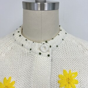K&ouml;nnte beinhalten: Wei&szlig;er Strickcardigan mit Knopfleiste und gestickten gelben Blumen. Der Cardigan hat einen gr&uuml;nen Punktensaum am Ausschnitt.