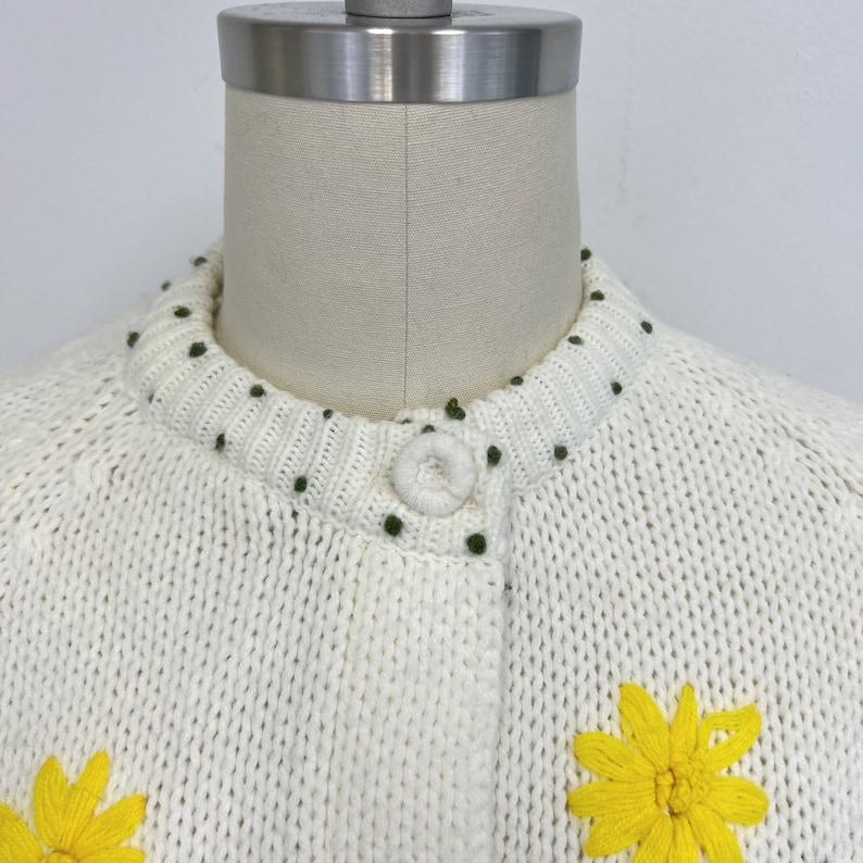 K&ouml;nnte beinhalten: Wei&szlig;er Strickcardigan mit Knopfleiste und gestickten gelben Blumen. Der Cardigan hat einen gr&uuml;nen Punktensaum am Ausschnitt.