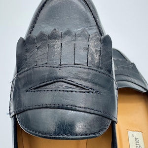 May include: Close-up of a pair of black leather loafers. The shoes feature a decorative fringe detail across the vamp and a stitched design. The interior sole is a light tan color.