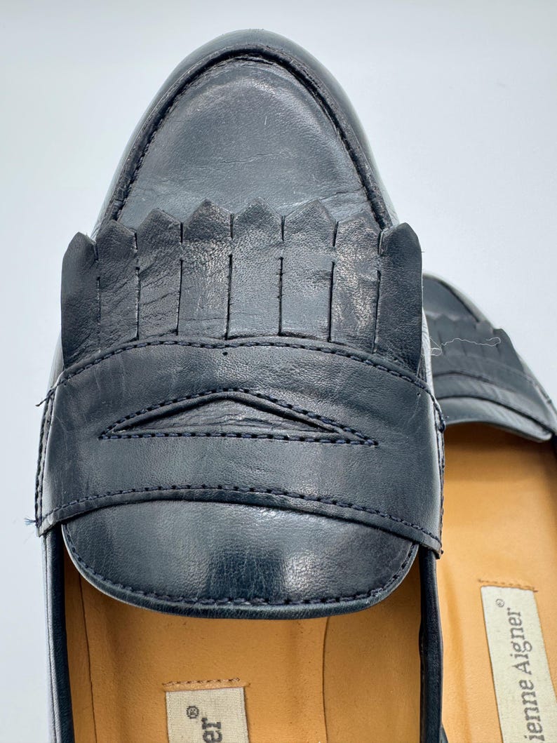 May include: Close-up of a pair of black leather loafers. The shoes feature a decorative fringe detail across the vamp and a stitched design. The interior sole is a light tan color.