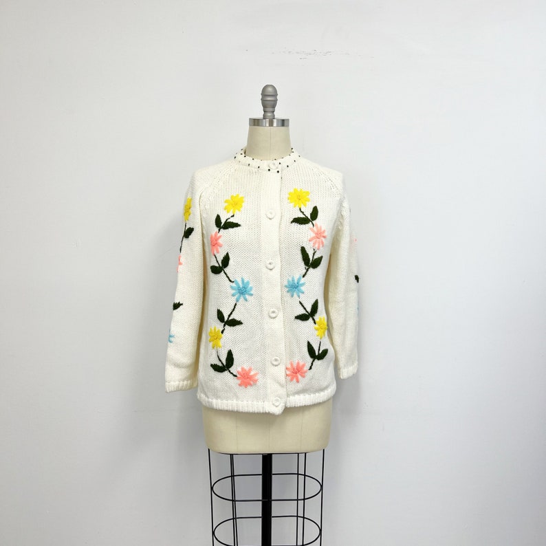 K&ouml;nnte beinhalten: Ein wei&szlig;er Strickcardigan mit Blumenmuster. Die Blumen sind in Gelb, Rosa, Blau und Gr&uuml;n gestickt.