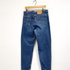 May include: Blue denim jeans hanging on a wooden hanger. The jeans have a classic five-pocket design, with a leather patch on the back. The jeans are a medium wash and appear to be vintage.