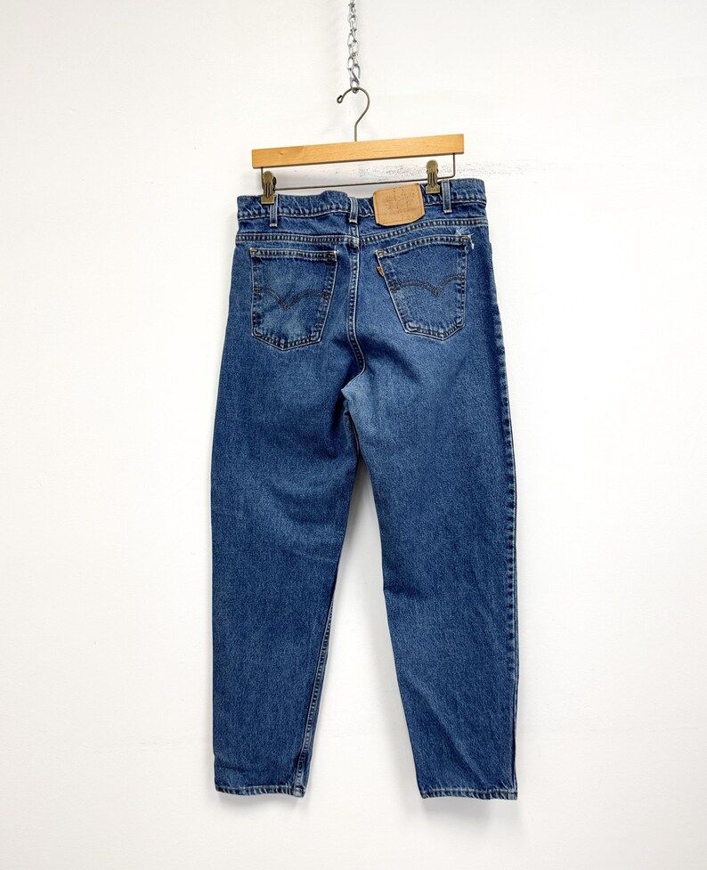 May include: Blue denim jeans hanging on a wooden hanger. The jeans have a classic five-pocket design, with a leather patch on the back. The jeans are a medium wash and appear to be vintage.