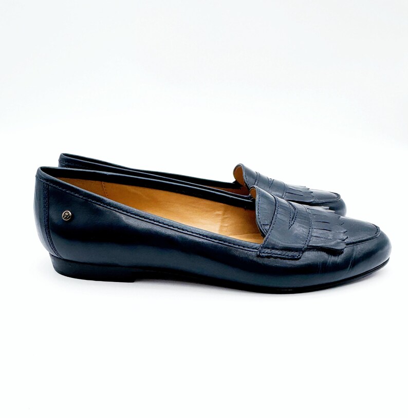 May include: A pair of navy blue leather loafers with a classic design. The shoes feature a rounded toe, a fringed vamp, and a small heel. The loafers are set against a white background.