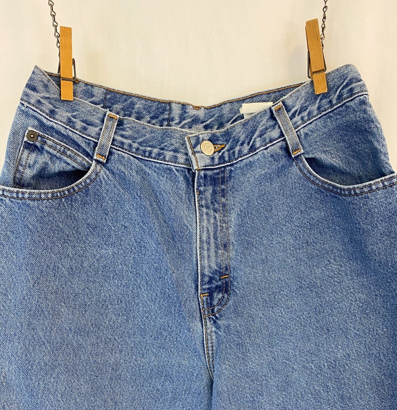 May include: Light blue denim jeans with a button closure and front pockets. The jeans are hanging from wooden clothespins. The denim has a classic, faded look.