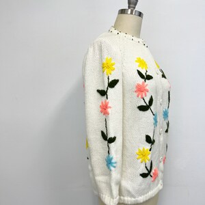 K&ouml;nnte beinhalten: Wei&szlig;er Strickcardigan mit gestickten Blumen in Rosa, Gelb und Blau. Die Blumen sind vertikal auf den &Auml;rmeln und der Vorderseite des Cardigans angeordnet.