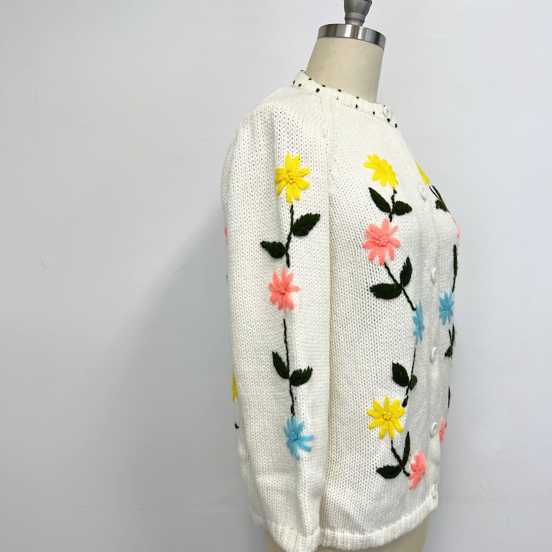 K&ouml;nnte beinhalten: Wei&szlig;er Strickcardigan mit gestickten Blumen in Rosa, Gelb und Blau. Die Blumen sind vertikal auf den &Auml;rmeln und der Vorderseite des Cardigans angeordnet.