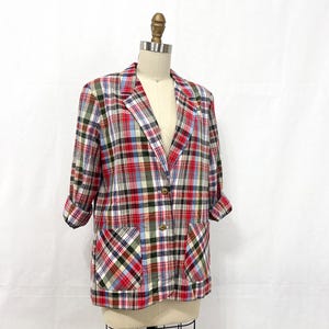 Può includere: Un blazer a quadri multicolore rosso, blu, verde e bianco, esposto su un manichino. Il blazer ha due tasche anteriori, maniche arrotolate e due bottoni color oro. Il tessuto presenta un classico motivo a quadri.