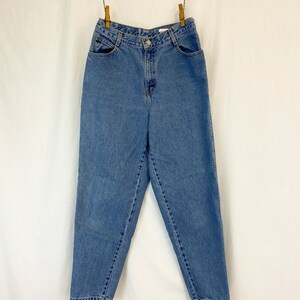 May include: Light blue denim jeans hanging from a clothesline. The jeans have a high waist, pockets, and a button closure. The denim has a faded appearance, suggesting wear. The jeans are suspended against a white backdrop.
