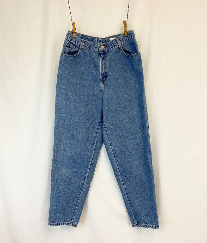 May include: Light blue denim jeans hanging from a clothesline. The jeans have a high waist, pockets, and a button closure. The denim has a faded appearance, suggesting wear. The jeans are suspended against a white backdrop.