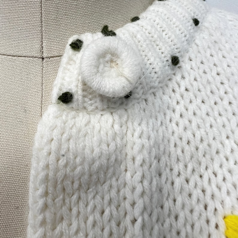 K&ouml;nnte beinhalten: Wei&szlig;er Strickcardigan mit einem gro&szlig;en wei&szlig;en Knopf und gr&uuml;nen Verzierungen. Der Cardigan hat ein Zopfmuster.