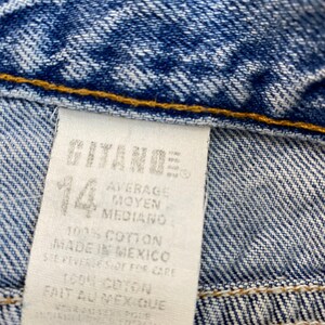May include: Close-up of blue denim fabric with a white tag. The tag reads "GITANO" and "14", with text in English, French, and Spanish. The fabric is a medium wash denim with visible stitching.
