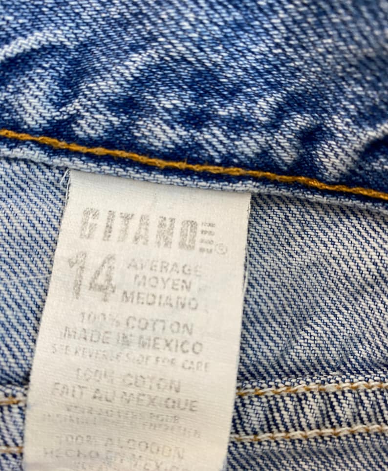 May include: Close-up of blue denim fabric with a white tag. The tag reads "GITANO" and "14", with text in English, French, and Spanish. The fabric is a medium wash denim with visible stitching.