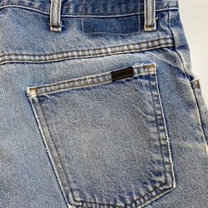 Pu&ograve; includere: Un paio di jeans blu denim con una tasca posteriore e una toppa in pelle marrone. I jeans hanno un lavaggio chiaro e un aspetto sbiadito.