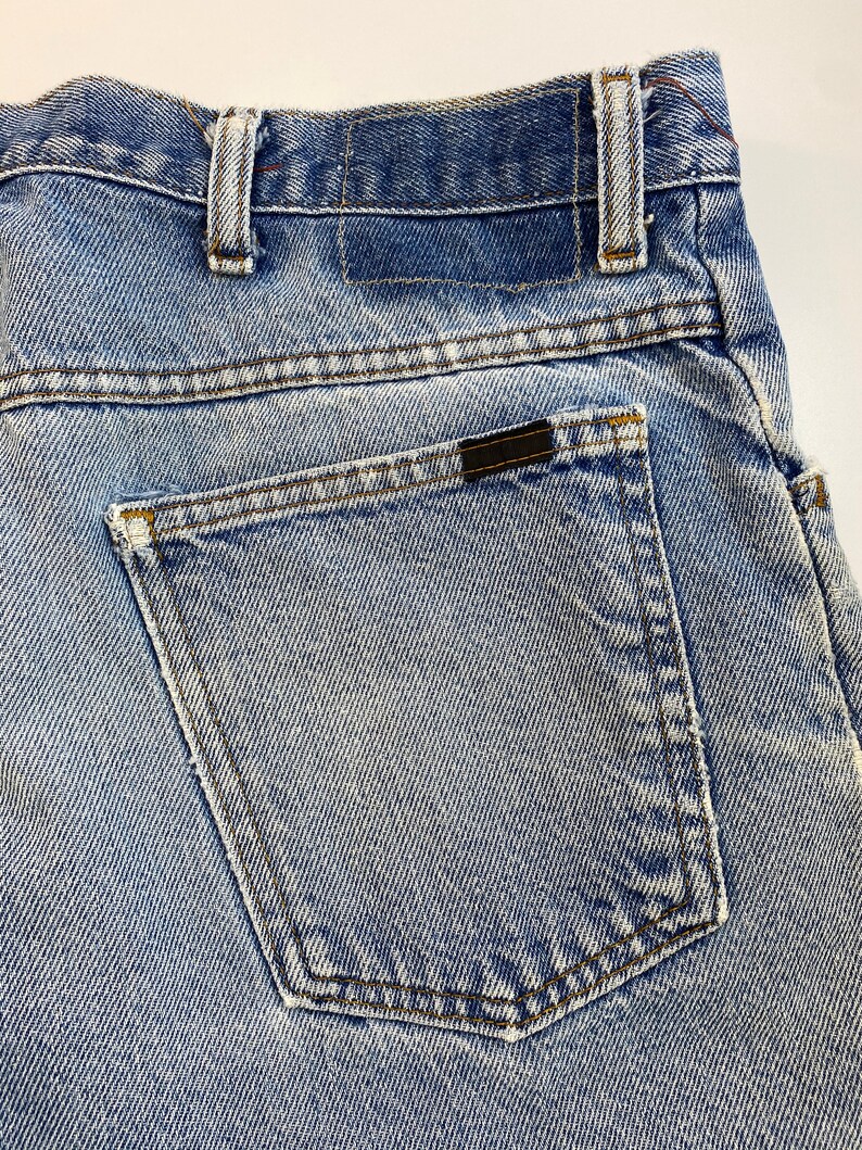 Pu&ograve; includere: Un paio di jeans blu denim con una tasca posteriore e una toppa in pelle marrone. I jeans hanno un lavaggio chiaro e un aspetto sbiadito.