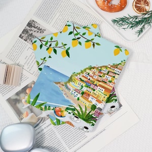 May include: Colorful art prints featuring a coastal town scene and lemon branches. The prints are layered on top of a newspaper, with a white plate and a pastry in the background. The art style is illustrative.