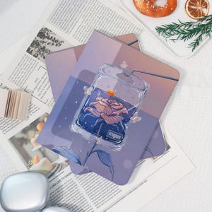 May include: A set of purple and lavender notebooks with a floral design. The notebooks feature an illustration of a rose inside a clear container with butterflies. The background includes newspaper clippings and a white surface.