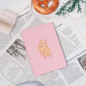 May include: A light pink tablet case featuring a watercolor illustration of a hamster giving a peace sign. The hamster is tan with a white belly. The case is on a newspaper background.
