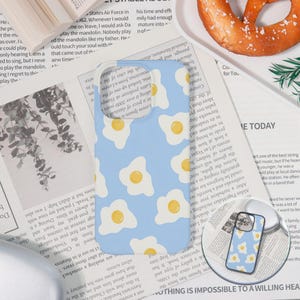 May include: Light blue phone case with a pattern of fried eggs. The case is shown on a newspaper background, with a pretzel and greenery in the background.